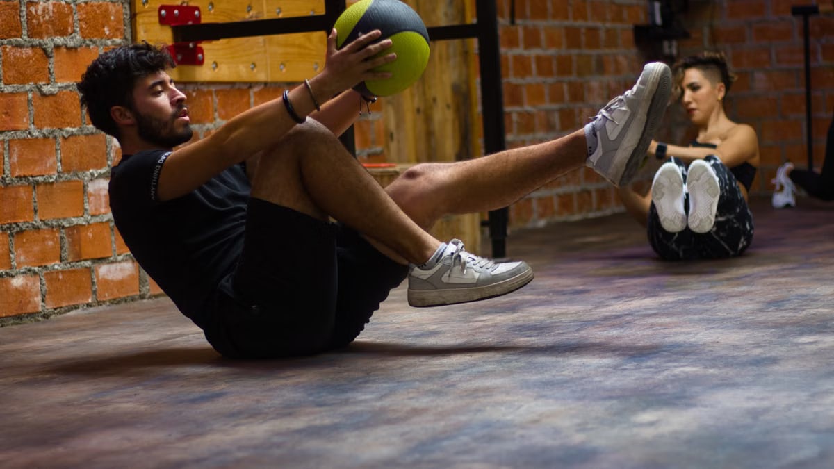 A man and woman perform core exercises with a medicine ball in a brick wall gym.
